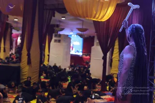 A photograph of a yoga session hall with yellow and maroon draped fabric canopies. In the right foreground a long-haired man in a leopard-print sleeveless top and maroon lower cloth stands with his back partly to the camera, raising a silver-and-white ceremonial staff overhead. Before him, a large crowd of participants in dark clothes sits and lies on colourful mats across the floor. A screen at the back shows a stage. The figure appears to be a person with long hair and distinctive ceremonial dress, though appearance is partly obscured.