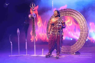 A man stands on a stage before a golden ornate circular arch and red-orange flame backdrop with blue haze. He wears a leopard-print cloth wrap over the chest, a beaded chest adornment, a rudraksha-style bead necklace, bangles, and maroon dhoti-style drapes. He holds a silver trident with a small damaru drum. Ceremonial props are arranged to his left, including a staff with a curved top, a dark feathered staff, and a silver rod with a drum. He has a red tilak on his forehead and a long braid, and stands on a patterned mat. The ceremonial costume suggests a Shiva-themed staged performance or darshan.