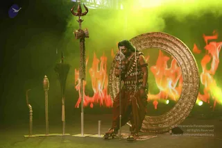 A man in a staged setting stands beside a large ornate golden circular frame, holding a tall silver trident with a damaru drum near its top. He wears a leopard-patterned sash, a cross-body garland of rudraksha beads, bracelets, and rust-brown dhoti-style cloth, with bare feet on a patterned mat. Behind him stand orange flames against a green-lit backdrop. Other ceremonial weapons and staffs, including a silver staff, a pointed rod and a dark ceremonial sword-like object with a tassel, are arranged on the left.
