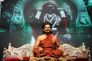 A seated man in an orange-red saffron wrap and dhoti sits cross-legged on an ornate silver-carved throne with red cushions, draped with a tiger-skin rug. He wears a white tilak on his forehead, rudraksha-style beaded necklaces, and dark braided hair, and holds his hands open, smiling while speaking into a microphone on a stand. Behind him a large green-toned screen shows a deity murti adorned with white flower garlands, a heavy white bead necklace, and red garland accents, framed by carved stone arches with lion motifs. A small red-and-white object sits on a silver tray at lower left. a person: long dark braided hair, saffron wrap, rudraksha-style beads and ceremonial necklaces, seated on the throne as the speaker.