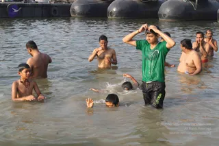 Several people bathe waist-deep in a river with murky greenish water, with black floating buoys or pontoons visible at the top of the frame. A young man in a green t-shirt and black shorts stands in the center, pushing wet hair back with both hands. Beside him a bare-chested man rubs his face, a shirtless boy in the foreground wears a red thread necklace, and other men and boys stand or swim nearby, some partially submerged.