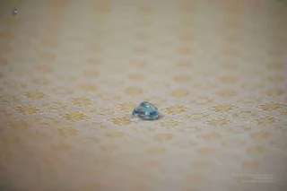 Close-up photograph of a single faceted pale blue gemstone, roughly pear or teardrop shaped, resting on a cream fabric with a repeating pattern of small golden-yellow floral motifs arranged in diamond-shaped grids. The background is softly blurred and shallow depth of field keeps the stone in focus. No people are visible.