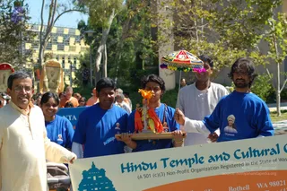 Outdoor procession on a sunny day with trees and a building behind. In the foreground a bearded man in cream kurta stands at left beside a woman in a blue t-shirt. Center, a young man in a blue t-shirt with a small printed portrait holds a wooden platform bearing a small figure draped in orange and marigold garlands, while a man in white kurta holds a small multicoloured ceremonial parasol above it. At right, a man in blue t-shirt with a printed portrait holds the parasol pole. A banner reads Hindu Temple and Cultural Center, Bothell WA. Several other people stand in the background.
