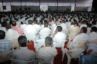 A large tented indoor gathering seen from behind the audience. Hundreds of men sit on white plastic chairs on a red carpet, many wearing white shirts and dhotis, facing a stage area out of frame. Some wear pale blue, grey and pink shirts. Banners hang on the tent walls and overhead lights are visible. The scene suggests a convention session with a seated congregation listening to a speaker.