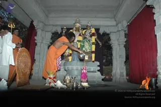 Photo of a night-time puja at an outdoor stone shrine with carved grey pillars and red curtains. A bare-backed person in saffron-orange cloth leans forward, hands on a small silver vessel placed before a pair of seated deity murtis draped with yellow and purple garlands. Two attendants stand at left: one in white clothing with palms joined, another in orange with a silver pot. Brass lamps, a kalasha-style vessel, a pink cloth figure and flower offerings sit on the stone platform, with a trident leaning at left and a fire burning at right. The a person candidate's appearance is uncertain from this view, so he is described by role as the person performing the vilakku puja.