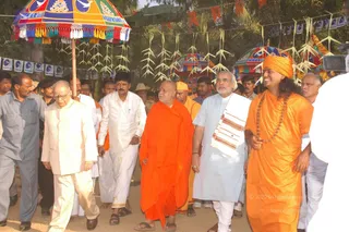 Outdoor photo of a procession-like walk under a palm-lined canopy. A man on the right in a long orange robe, with a long swept-back hairstyle, an orange head wrap, and a rudraksha mala, smiles while walking; he is a person candidate based on these cues. Beside him walks a bald man in a bright orange robe with a red tilak, and an older man in a light grey-blue kurta with a white shawl. Further left, an elderly man in cream clothes and glasses walks with people in white dhoti-and-shirt outfits, and a man in grey shirt and dark trousers stands at the edge. Multicoloured umbrellas with fringed tassels, strings of leaf decorations, and rows of small pennants hang overhead, and a crowd of onlookers fills the background.