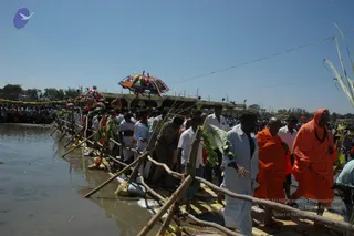 Outdoor photograph of a riverbank ritual procession at a water edge, with a line of men in white dhotis and shirts walking along a bamboo-and-banana-leaf fence at the shoreline. Two figures in saffron-orange robes stand prominently in the foreground on the right; the front figure wears an orange hooded wrap and dark beads and walks with a hand gesture, while the other wears a saffron robe. Colourful striped and patterned ceremonial umbrellas are raised above the procession, and a crowd of onlookers gathers behind a yellow-decorated pavilion under a clear blue sky.