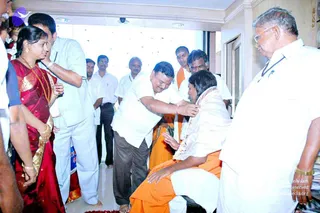 A photo inside a bright showroom with white walls and a glossy floor. In the centre, a man in a white shirt and grey trousers bends forward to place a garland over the head of a seated man in white clothes with an orange saffron shawl and a garland of flowers, who is seated low in a chair-like pose with hands near the floor. Nearby, a woman in a red and gold silk sari with gold jewellery and bangles stands at left, and a man in a white shirt stands beside her. A man in an orange shawl stands behind, and a man in white with rimmed glasses and a gold wristwatch stands at the right edge. Several other men in white shirts and dark trousers stand in the background near the entrance. Petals are scattered on the floor and a glass-fronted display area is visible behind them.