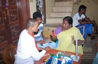 A photograph of a medical camp at an outdoor doorway. A man with a shaved head, wearing a white coat and a red scarf with a rudraksha-style necklace, sits at a small wooden table across from a woman in a yellow saree with a purple blouse and gold bangles, who gestures with one hand while holding a small orange pouch. A young man in a white t-shirt with a lanyard sits beside them writing on a clipboard, and another young man in a white shirt and blue striped track pants sits on the steps behind. The table holds blister packs of tablets, a blood pressure cuff, a laptop, a stethoscope and medicine boxes, including a Lacto-Calamine box. Wooden double doors with carved medallions stand to the left.