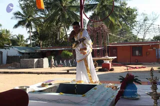Outdoor ritual scene at a temple-like compound under palm trees. A man in a white dhoti with gold-bordered cloth and a gold-bordered shawl stands at a square stone fire pit or yagna kund, pouring from a metal vessel. Brass oil lamps, a blue pot with red flower garland, a coconut, and yellow marigold petals rest on the platform. Orange-painted buildings and a red metal frame structure stand behind, with a small figure walking in the background and rows of small stone figures on the left. The scene is a homa-style ritual at an ashram setting.