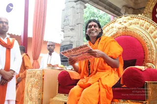 A smiling person with long dark hair and a red bindi sits on an ornate golden throne with red cushions, wearing a saffron-orange robe and a dark rudraksha mala. He holds up a printed sheet with red and black text. To the left, a man in a white shirt with an orange shawl stands beside another man in saffron clothing. A carved stone pillar and a striped red-and-orange drape are behind the throne. A small brass tray with offerings sits at the side.