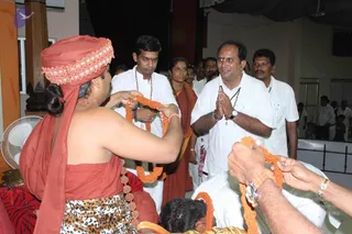A photograph of a devotional greeting at an indoor hall. A figure seated at left, wearing a rust-red turban with a decorative band, a rust-red cloth wrap and a leopard-print lower garment, holds up a long orange garland being offered by hand. Behind them, a man in white shirt and dhoti with a rudraksha mala stands beside a woman in rust-red sari with a rudraksha mala, and a man in white with hands pressed together in namaste smiles. Several other men in white shirts stand in the background, and an orange garland is held by hands at lower right. A red-patterned cushion and a fan are visible at left, with a wooden pillar and an orange-and-white backdrop.