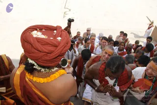 Outdoor photo on pale stone steps leading down to a river ghat. In the foreground, a person seen from behind wears a large red-orange turban decorated with white flowers, an orange-and-gold shawl, a marigold garland and a rudraksha-style beaded necklace, and holds a small metal vessel. Facing them, a group of devotees in white clothes and red-orange shawls with green lanyards stands on the steps, several with orange tilak on their foreheads. A man in the centre with a bare upper body and red shawl bows with palms pressed together. A cameraperson with a camera is visible at the top, and a figure in white and orange stands far up the steps at the right.