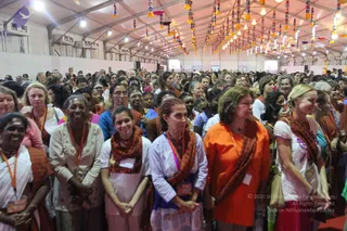 A large crowd of women fills a white tented hall decorated with hanging tassel streamers and strings of lights under the canopy. In the foreground, women in white, cream and pastel tops and orange or rust-coloured shawls stand closely together, many wearing orange and white lanyard badges. A woman in a bright orange top and rust shawl stands centre-right, and a woman in a white embroidered top with a pink-and-white skirt stands at the right edge. Air-conditioning units line the tent walls behind the crowd. No identifiable a person figure is visible in this image.
