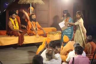 A stage event at dusk with a seated group of saffron-clad sannyasis on an orange-cushioned dais under a decorative parasol. On the left, a man in a dark red-brown robe with a yellow marigold garland and gold armlets smiles and gestures, wearing a black turban. Beside him, a bearded man in orange robes with a rudraksha mala and a red forehead mark sits cross-legged, with an attendant holding the parasol behind them. On the right, a woman in a blue patterned sari wears a black blindfold and holds a microphone and a clipboard, while another woman in a cream sari with a pink border gestures toward her. Seated devotees in orange and white clothing with folded hands watch from the front, and a man in orange with a turban sits near the audience. The backdrop is a wooden panelled wall.