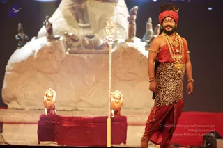 A man stands on a stage beside a gold-tipped silver trident, in front of a large pale stone sculpture of a seated figure with a carved Shiva lingam-like mass and several smaller deity murtis. He wears a red turban, rudraksha-style gold and beaded necklaces, gold armbands and bracelets, a red draped garment, and a leopard-print lower wrap. Two golden lion heads flank a red velvet-draped platform in the foreground. Swamiji (probable) cues: long swept-back hair under a red turban, ornate gold necklaces and saffron-red ceremonial clothing; the figure is standing on stage in an apparently presentational pose.