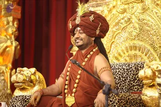 A man sits on an ornate golden throne with a leopard-print cushion, smiling and speaking into a microphone on a stand. He wears a rust-red turban decorated with gold ornaments and a white feather, a rust-red sleeveless wrap, a long gold bead necklace with a gold pendant, and red bangles on his wrists and upper arm. Gold lion-head armrests and a red curtain frame the scene. The turban, red-and-gold clothing and throne match the presentation of Swamiji (probable) seated for satsang. A printed copyright line appears at the lower right of the photo.
