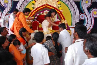 A photograph of a devotional event at a decorated throne canopy with an ornate gold and red throne backdrop garlanded with yellow and red flowers. A seated figure with long swept-back dark hair wears a white and gold-bordered garment, multiple gold necklaces and bangles, and reaches down to a devotee. A man in an orange-saffron robe stands at left holding a tray or offering, while a man in white stands at the left edge. Around the throne, a crowd of devotees in white shirts, orange and saffron saris and a grey-haired woman presses forward; one woman holds a small child in light clothing and another woman with a long braid in a dark patterned sari holds a fruit offering. Swamiji (probable): the seated figure with long swept-back hair, gold necklaces and a white-and-gold garment, seated on the throne, matches the page's Swamiji-at-event note. Several dozen people are visible.