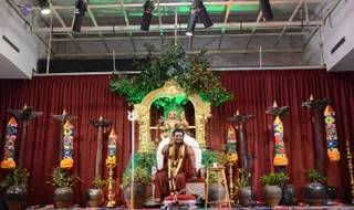 A seated man in rust-brown robes sits on a red-cushioned throne on a stage before a deep red curtain. He wears heavy gold necklaces and a rudraksha mala, and raises one hand in a blessing gesture. Behind him a gilded arch frames a golden deity murti of a seated figure with a child, set among green foliage lit with green light. Tall wooden poles with striped colourful fabric banners flank the stage, with brass lamps, potted plants, a trident and a small stool with a cup in front. Stage lights hang from a metal truss overhead. The man is wearing a red garment and gold necklaces, consistent with a satsang or discourse setting; a probable match to a person appears in the appearance-based cues.