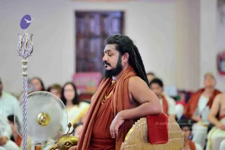 A seated man with long dark dreadlocks and a full beard wears a saffron-orange wrap over his shoulders and a gold necklace, with eyes closed, resting his arm on a gold-trimmed throne with a red cloth draped over it. In the foreground a silver trident stands on a pole topped with a purple emblem, beside a brass lamp base. Seated devotees in white and orange clothing fill a hall behind him, and a pink banner hangs on the wall.