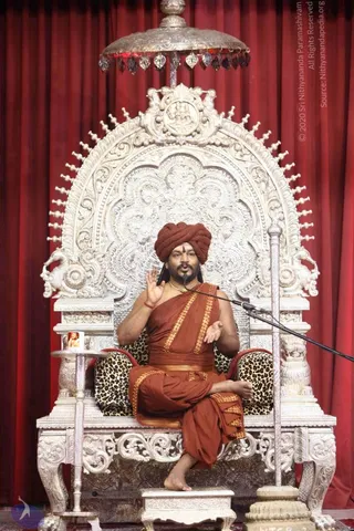 A man in a rust-orange turban and matching orange robe sits cross-legged on an ornate silver throne with carved floral and scrollwork backrest, a silver canopy with hanging crystal drops above, and leopard-print cushions. He raises one hand while speaking into a microphone on a stand. A small framed portrait sits on a silver side table to his left. Behind him hangs a deep red curtain. A rudraksha-style necklace and a red thread on his wrist are visible, with a tilak on his forehead. Swamiji (probable): he wears a turban, saffron-orange robe and ornate gold necklace, consistent with the page's reference to him teaching at the event.