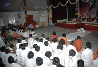 A seated group inside a covered hall with a red-and-cream stage at the right. Many devotees in white clothes sit closely on the floor, mostly facing away from the camera, with several in saffron/orange robes among them. Some sit on white mats and a cushioned mat in the foreground. Behind them a small easel with a blank white board, potted plants, and colourful paper garlands hang from the ceiling. On the upper right a large framed poster shows a close-up face portrait in red tones. A small group of saffron-clad figures sits on the raised stage in the background.