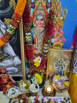 A home shrine photographed close up, with no people visible. Central is a framed deity murti of a goddess in red and gold ornate garments, heavily jeweled with layered necklaces and a crown, shown against a red and gold halo. She is draped with a thick garland of red and orange marigolds and white and yellow flowers. To the left is a black-and-white photograph of a seated figure with bare feet and a bracelet of beads, partly cropped. Lower left, a small figure in an orange turban and orange robe with gold necklace sits in a printed image. To the right, a gold-framed picture shows a standing deity with a tall crown and blue skin in an arched shrine. In front are offerings: a glass bowl with a banana and cut fruit, small bowls of rice and liquid with jasmine flowers, a lit oil lamp in a brass holder, and a bowl of purple and yellow flowers. A tray of flowers and dishes lies in the foreground.