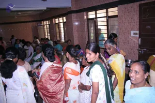 A crowded indoor dining hall with pink tiled walls and wooden-framed windows. Many women in saris stand and move in a queue near a counter holding steel vessels and serving pots. In the foreground, a young woman in a white sari with green border and a jasmine-braided plait smiles toward the camera. Beside her, an older woman in yellow and white sari with glasses holds a plate. A woman in a red and gold sari stands at left holding a small container, and another in an orange-bordered white sari stands nearby. No a person candidate is visible; the scene shows volunteers and devotees at a meal service.