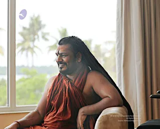 A seated man with long dark hair swept back, a red-orange ochre wrap draped over one shoulder and bare chest, and a red tilak mark on his forehead, smiling while looking to his left. He rests one arm on a beige upholstered armchair. Behind him a large window shows palm trees and greenery, and a cream curtain hangs at right. Swamiji (probable) is visible here, shown by the long swept-back hair, orange ochre wrap and distinctive forehead marking; in_person applies as he is physically present in the room.