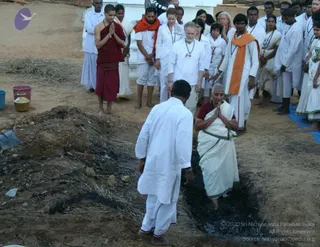 A photograph of a firewalk ceremony at an outdoor ashram site with dry ground. In the foreground a man in white dhoti and kurta stands at the edge of a dark, charred pit of embers, and a grey-haired woman in a white sari with a maroon blouse stands in the pit, hands in anjali mudra. Behind them a crowd of devotees in white clothing, some with orange shawls and rudraksha malas, stands with hands folded. A saffron-robed figure in maroon wraps and a brass kalasha or pot sit to the left. A blue tarp lies at the right edge.
