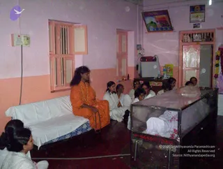 Indoor photo of a small room with pink-and-white walls. A long-haired man in saffron-orange robes with a beaded mala necklace sits cross-legged on a white-draped sofa at left, hands resting in his lap. Beside him and along the wall sit several women and girls in white clothing, seated on the floor. At right stands a glass-sided wooden casket holding a body wrapped in white cloth and scattered with flower petals, with a garland hung on the wall nearby. Framed pictures hang above the doorway, and a small red radio and water bottle sit on a shelf. Seated figures in the frame total about eight people. Swamiji (probable): long hair, saffron robes and rudraksha-style mala are visible on the central seated figure, labelled by the page as the Swamiji present at this event.