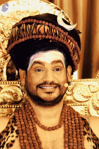 A close-up photograph of a smiling man seated before an ornate gold carved throne with a curtain behind him. He wears a tall black turban wrapped with a tiger-striped band strung with rudraksha beads, and a crescent moon ornament is tucked at the side. A white vertical forehead mark is applied, a small lapel microphone is clipped near his face, and he wears hoop earrings. Several strands of rudraksha malas hang over a bare chest, with a leopard-print shawl at the shoulders.