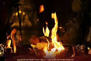 Close view of a sacred fire burning in a ritual fire pit during a homa. Tall orange flames rise from a mound of offerings, including a pale block, a reddish-brown rounded item, and grains or seeds scattered on dark wood logs. A brass or golden ritual vessel sits at the lower left edge, and a blurred golden ornamental backdrop with lamp-like lights fills the dark background. No people are visible in the frame.