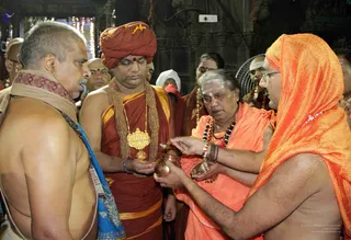 A photograph inside a temple with warm lamplight and string lights in the background. A bare-chested priest in a white dhoti with a blue-bordered shawl and a lanyard stands at left. Beside him, a man in a rust-red turban, rudraksha mala and a large ornate gold pendant holds a copper pot. Next to him an older man in orange robes, rudraksha malas and forehead tilak pours liquid from a small cup into the copper vessel. At right, a bare-chested man in an orange head-cloth and orange cloth holds the pot, with other devotees and attendants visible behind. The scene resembles an abhishekam offering at a temple shrine.