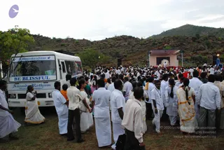 Outdoor gathering on a grassy hillside below rocky boulders and a green hill. A large crowd of devotees, mostly men in white dhotis and white shirts with some women in white and cream sarees with gold borders, stands close together facing a raised stage at right. The stage has a pink-roofed canopy, a backdrop with a framed image, and a red cloth. At left, a white bus with a blue-lettered sign reading Nithyanandam and BE BLISSFUL, and Ananda Ratham Nithyanandapeetam painted on its side, stands on the grass beside the crowd.