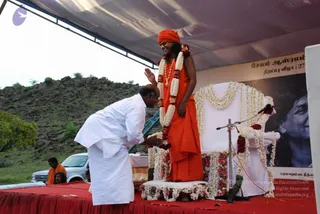 A figure in an orange-saffron robe and orange turban stands on a flower-decorated white throne on a red-draped stage under a tarpaulin canopy. He wears a white flower garland and an ornate gold necklace, and raises one hand in greeting. A man in a white shirt and white dhoti bends forward to offer his hands in reverence. A banner beside the stage shows a large portrait labelled "Paramahamsa Nithya..." and Tamil text. Rocky hills and a parked white vehicle are in the background, and a seated figure in orange appears at the left edge. Visible cues are consistent with Swamiji (probable) on the throne, given the orange robe, turban, garland and gold necklace.