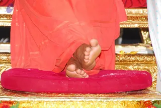 Close-up photo of a seated figure's bare feet resting on a round red velvet cushion placed on an ornate gold-carved throne platform. The figure wears orange-red robes that drape over the seat. Red and yellow flower petals or offerings are visible at the bottom edge, and a white curtain with blue dots appears at the right. Only the feet and lower robes are visible, so no face or upper body is shown.