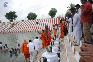 Outdoor photo of a ritual at a water tank with red-and-white striped steps. A long-haired figure in saffron-orange robes, wearing a red-and-white garland and rudraksha-style beads, stands at the water's edge with raised hands. Beside them a second person in orange holds a staff, and a bald man in orange robes wades in the water. Several people in white dhotis and kurtas stand on the steps and in the water, some with folded hands. Along the steps, black stone lingams draped in white cloth and garlanded with flowers sit in a row. Palm trees and a grey sky appear in the background. a person is the long-haired figure in saffron with the garland and beads.