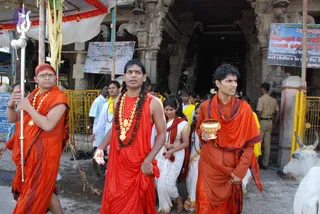 A procession at a temple entrance with a yellow-railed barrier and carved stone pillars. In the centre, a man with long dark hair, a rudraksha mala and a gold necklace wears a saffron-orange dhoti and shawl, walking with his palms open. On the left, a man in saffron robes and an orange head wrap with a rudraksha-bead garland holds a silver trident-staff. On the right, a curly-haired man in saffron robes carries a gold-coloured vessel. Behind them, women in white saris with red borders and men in white and yellow cloths follow. A white cow stands at right and a uniformed police officer stands in the background. Overhead hangs a white canopy with red trim, and signboards with Tamil and English text are on the walls.
