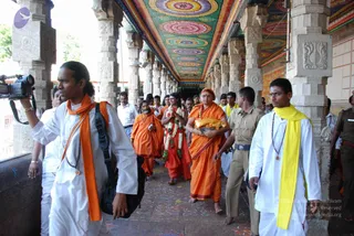 A photograph inside a temple colonnade with a richly painted ceiling of concentric circular patterns in red, green, yellow and blue, supported by carved stone pillars. A group walks along a patterned floor. In front, a man in white with an orange shawl and a camera-carrying companion; a central figure in saffron-orange robes holds a folded yellow cloth and a tray, wearing a large white flower garland; a bearded man in orange robes walks behind; a man in white with a yellow shawl and rudraksha mala walks at right beside a uniformed security officer in khaki. Several other people in white shirts and dhotis follow. a person: the central figure in saffron robes and a long garland, with cues of saffron clothing and ceremonial garland, depicted in procession through the temple.