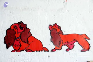 Two hand-cut red cartoon dogs are mounted on a white textured wall. The dog on the left sits and looks sideways with a purple collar, and the dog on the right stands in profile looking upward, also with a purple collar. Both have long, feathery reddish-brown ears and flowing fur, in a style resembling the animated characters Lady and the Tramp. No people are visible.