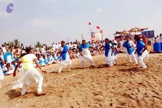 Outdoors on sandy ground, several women in white salwar-style outfits with blue shawls and one in yellow sash play a field game, running and reaching toward each other across chalk-marked lines. A large crowd of people in white and blue sits and stands along the edge of the sand. Behind them a stage with striped and colourful canopy tents, red and saffron flags, and a banner with a portrait stands under a clear sky. A few men in white and one figure in orange robes stand near the stage. A portrait in the banner is too small to identify.