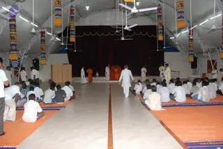 A photograph inside a large white tented hall with hanging decorative pillars patterned with elephants and motifs. Rows of seated devotees in white clothes sit on orange and blue mats facing a central aisle leading to a stage draped with dark red curtains. On the stage, a figure in orange robes sits on a red throne-like seat, flanked by standing people in white and yellow clothing. A man in a white kurta and dhoti walks down the central aisle toward the stage. Ceiling fans and lights hang overhead.