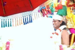 A smiling man sits under a red canopy hung with multicoloured tassels in blue, green, orange and pink, seated on a decorated palanquin or ratha. He wears a white turban-style headwrap with a gold border, a red garland of flowers around his neck, a red tilak on his forehead and bare shoulders with a white cloth. Golden finials and yellow and pink flower garlands decorate the canopy frame.