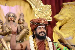 A bearded man in a maroon-brown turban with a leopard-print band and a small gold emblem sits before a microphone, right hand raised in a gesture while smiling. He wears a white jasmine garland, layered gold and beaded necklaces, a red and gold rudraksha-style bracelet, and a saffron-red wrap, with white vibhuti and a red kumkum dot on his forehead. Behind him stand gilded deity statues and an ornate golden lion-shaped throne armrest against a deep red curtain.