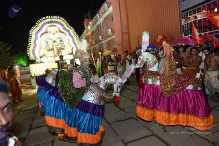 Night procession at an ashram with a brightly illuminated palanquin or float on the left, decorated with a large arched halo of white, green and purple flowers and lights, carrying a deity murti under a lit canopy. In the foreground two performers in elaborate costume ride or walk beside horse-shaped effigies with white and pink plumes, mirror-work and fringed red and green trappings; the man on the left wears a turban with a white feather, a green sequined bodice and a blue, purple and orange skirt, while the woman on the right wears a magenta and orange skirt with a red shawl and headdress. Behind them bare-chested men in white dhotis and orange shawls, some wearing rudraksha malas, follow with pink and red umbrellas and saffron flags. Lit building facades and string lights frame the scene.