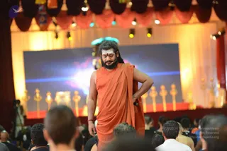 A bearded man with long swept-back dark hair stands on a stage in a saffron-orange draped robe, eyes closed, one hand resting on his hip and a red tilak on his forehead. A small headset microphone is visible near his face. Behind him a large screen shows a blue and pink glowing image, flanked by a row of gold candlestick-style lamps and gold-and-red stage drapes with warm lighting. A dense audience of many heads is seen in the foreground. The man appears to be Swamiji (probable), identified by long swept-back hair, saffron robe and stage presence, though his face is not used for identification.