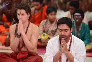 A photograph of a seated satsang gathering. In the foreground, a bare-chested man with long dreadlocked hair, a vertical tilak on his forehead, rudraksha mala and red dhoti sits with palms pressed together in anjali. Beside him, a man in a white kurta with a rudraksha mala and tilak sits with eyes closed, hands in anjali. Behind them, many seated devotees wear orange and saffron robes and white or green clothing, with a boy in a patterned shirt and a person in teal kurta visible. The scene suggests a devotional or meditative assembly. No identifiable a person is clearly present; the dreadlocked man's appearance is ambiguous.