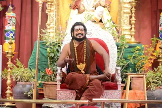 A photograph of a bearded man with long dark hair seated cross-legged on an ornate white and red throne, eyes closed and smiling, one hand raised in a gesture. He wears a rust-red and brown saffron-toned cloth, a rudraksha mala, a large gold ceremonial necklace with a central pendant, and a red tilak on his forehead. Potted green plants with a red flower surround the throne. Behind him stands a gold-clad deity murti with a golden kalasha and oil lamps on brass stands. A brass rod stands at the left. Probable Swamiji (probable): long hair, rudraksha mala, ornate gold necklace and saffron-toned clothing, seated on the throne. One seated person is visible.