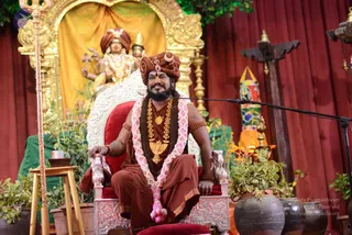 A seated man in rust-brown and saffron ceremonial robes sits on an ornate silver-and-red throne-style chair on a raised stage, wearing a rust-brown turban with a crescent emblem, a pink-and-white flower garland, a rudraksha mala, and gold necklaces. He has his right hand resting on the armrest and is speaking into a microphone on a boom stand. Behind him stands a gilded golden arch with a yellow drape and a deity murti in a turban and gold jewellery holding a trident-like staff. Potted green plants, brass lamps and a tall vertical decorative pillar surround the stage, with red curtains as the backdrop.